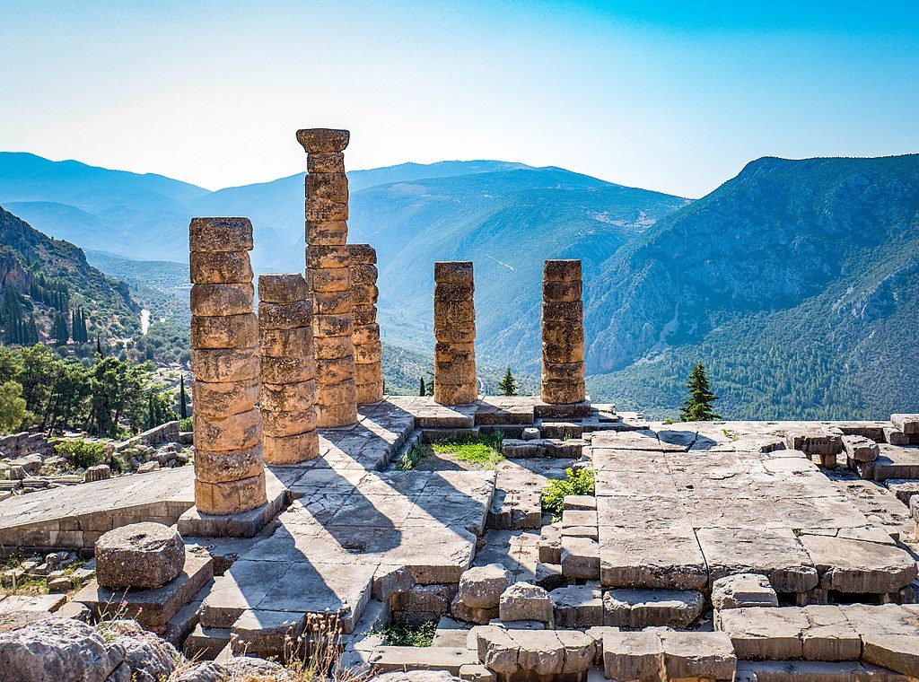 A picture of a beautiful landscape in Greece on a sunny day. In the foreground are ancient stone pillars from a temple. In the background are forest-covered mountains.