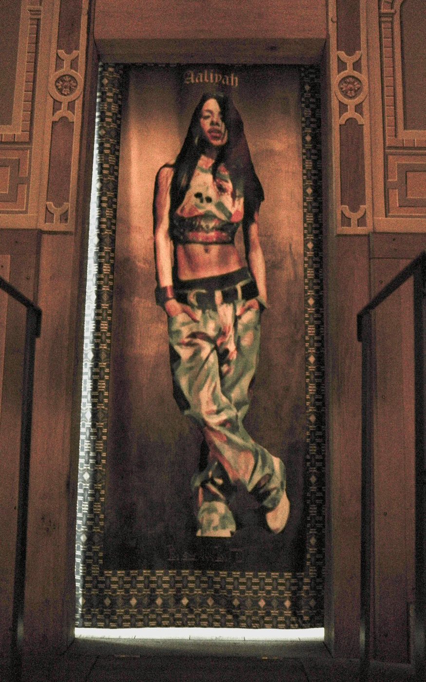 A tapestry of a person standing with their hands in their pockets, wearing a patterned crop top and jeans. They have long dark hair. The tapestry has patterned edges and hangs within a wooden doorframe.