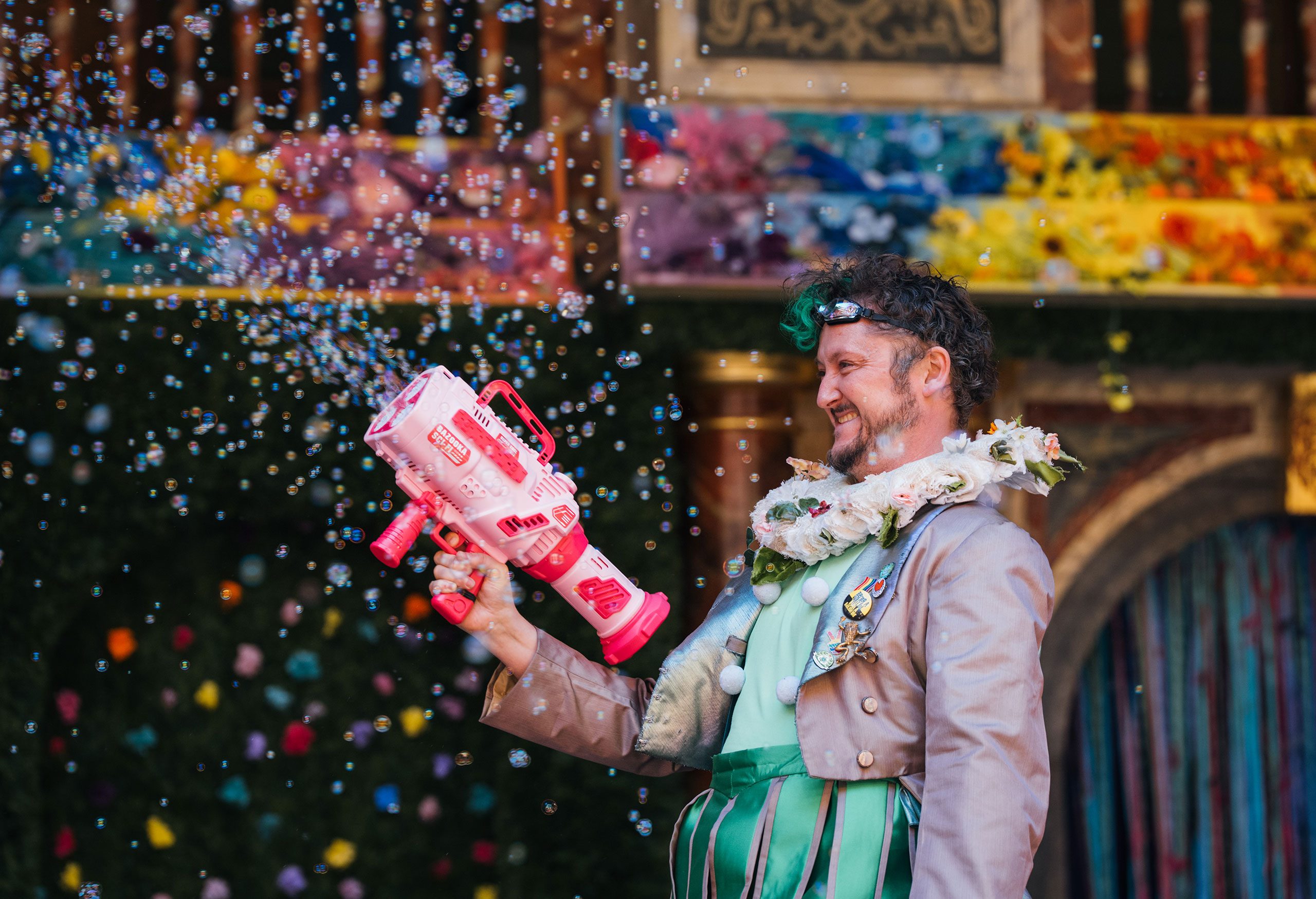 A white man with curly brown hair stands holding a pink bubble gun, spraying bubbles into the air. He wears a green costume and a white, flowery ruff.