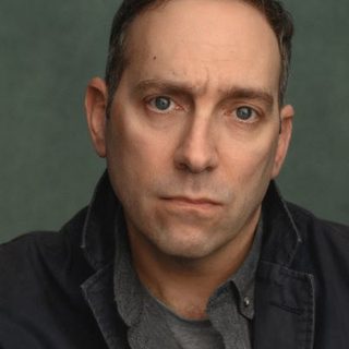 An actor with short grey hair and blue eyes looks into camera