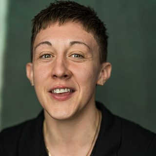 Charlie Josephine smiling directly at the camera. They are wearing a black shirt and have short dark hair.