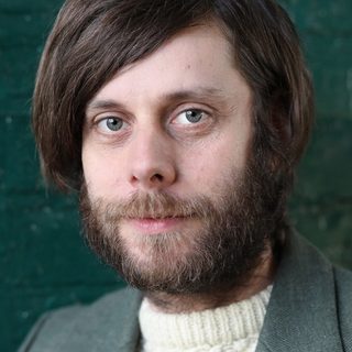Colin Michael Carmichael wearing a grey suit jacket over a cream jumper and looking directly at the camera.