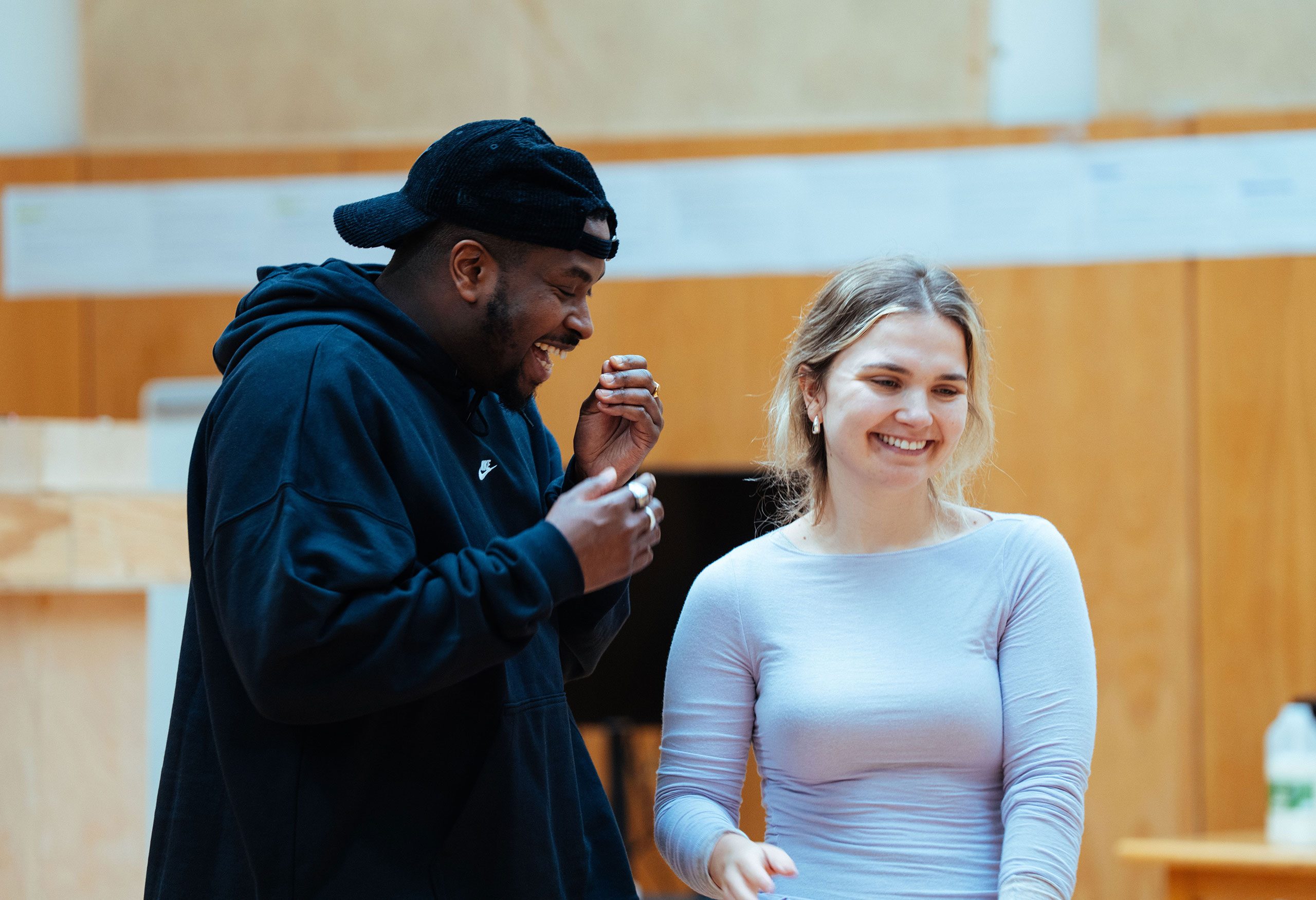 A British Gambian man wearing a black backwards cap and a white woman with blonde hair stand beside each other, laughing.
