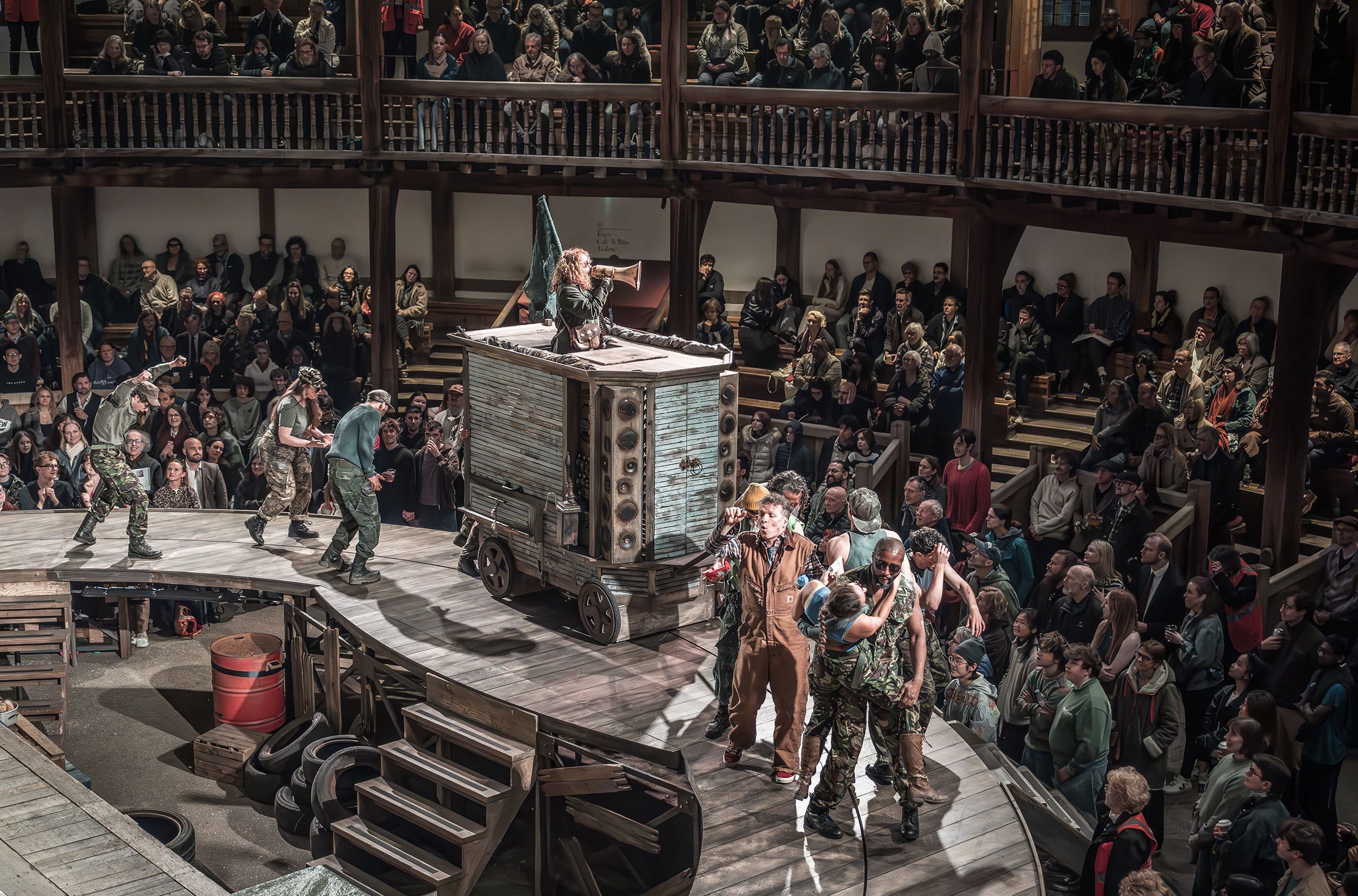 In a round theatre with wooden seating galleries, an audience watches as a parade of people wearing camouflage dance around a large blue cart. Standing in the cart is a white woman with long curly hair, speaking into a megaphone.