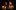 A woman and man stand in the glow of candlelight, the woman's hand touching the man's chin.