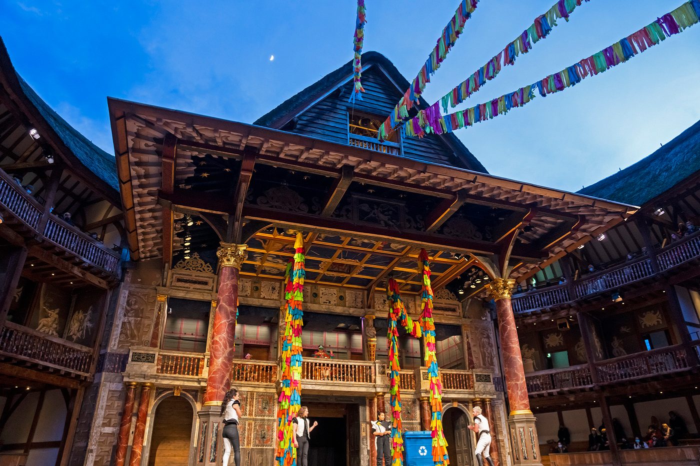 A twilight night sky, with a glimpse of the moon, shines above the Globe Theatre stage: with colourful streamings hanging across the open-air roof.