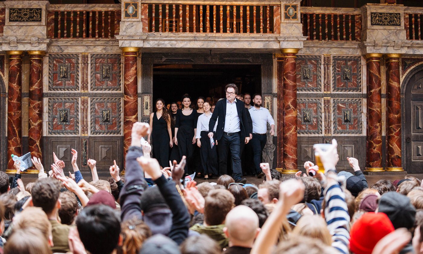 A crowd of people watch as a man leads a group of young actors on to a stage.