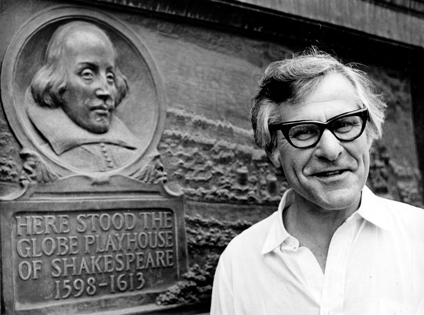A man wearing thick-framed glasses stands by a plaque marking the location of the original Globe Theatre