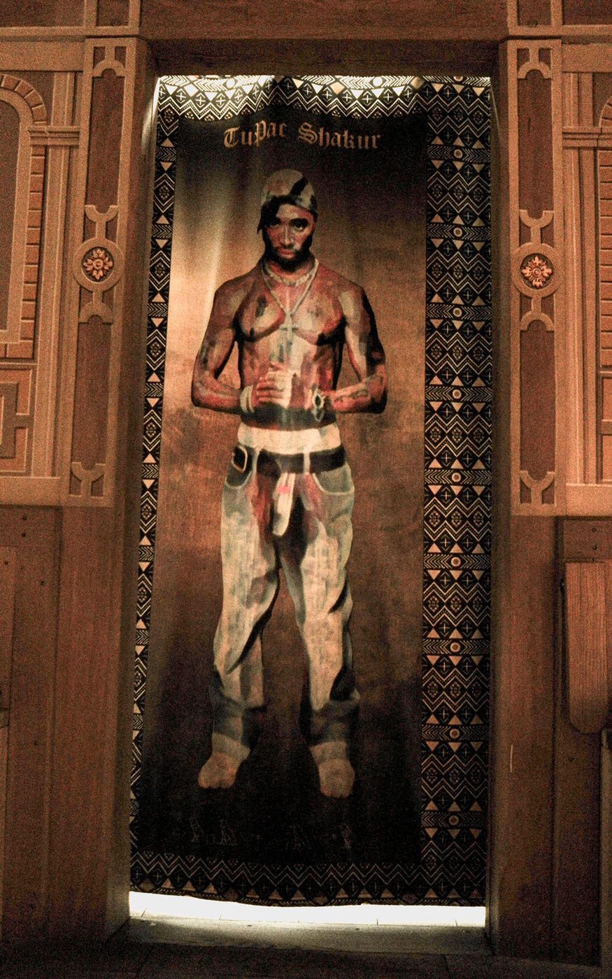 A tapestry of a person standing with their palms clasped together. They are shirtless and their chest is covered in tattoos. They wear a silver cross around their neck and baggy jeans. The tapestry has patterned edges and hangs within a wooden doorframe.
