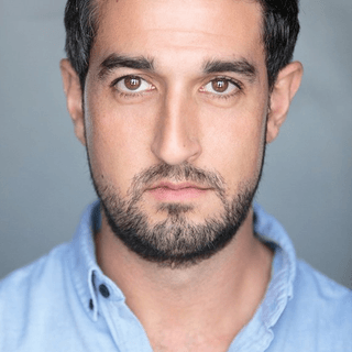 An actor looks into the camera. He has short dark hair and is wearing a blue shirt.