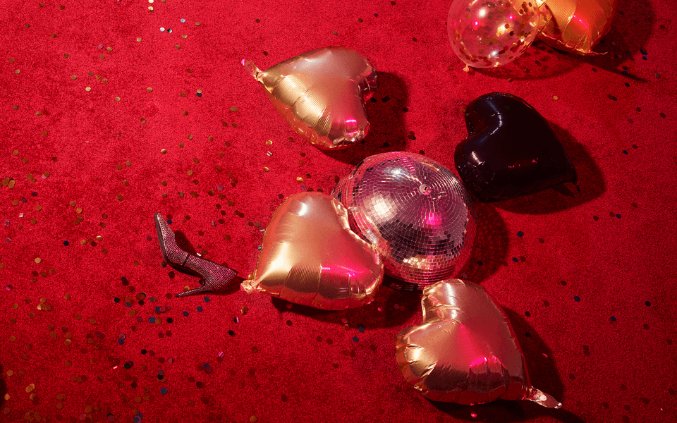 A red carpeted floor covered in glitter, heart-shaped balloons and a high heeled shoe.