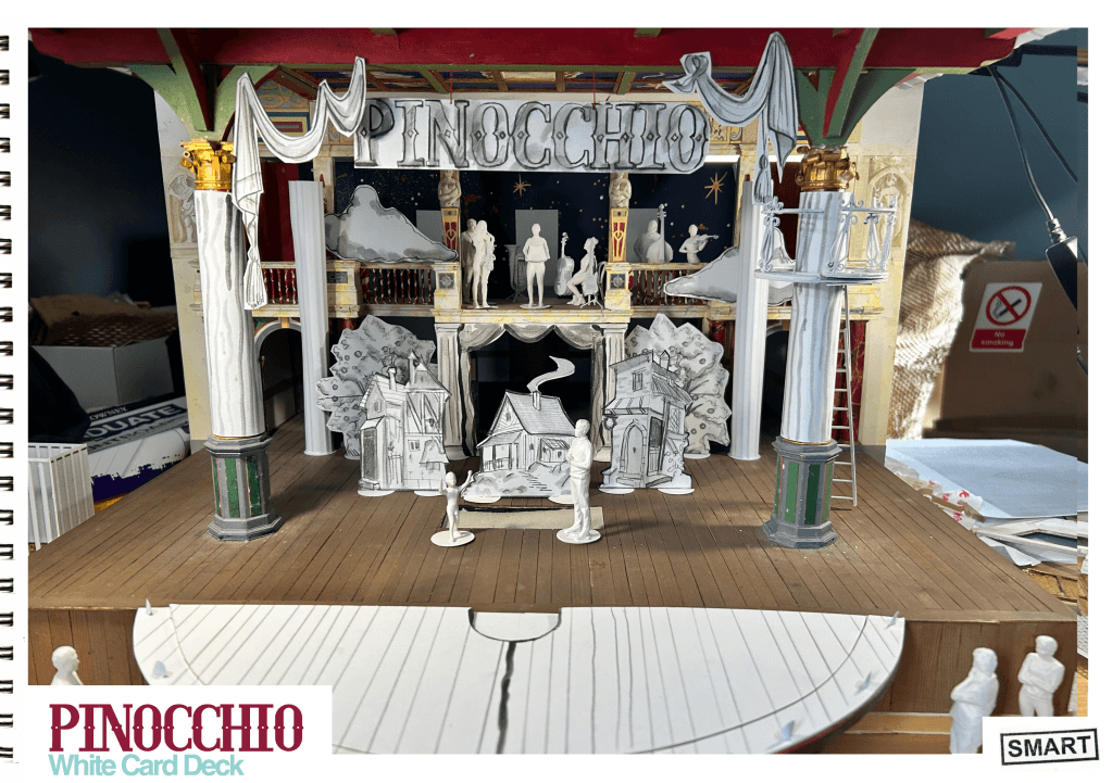 A model of the Globe Theatre. On the stage are pieces of set made of white paper, including houses, trees, curtains and a big sign saying 'Pinocchio'.
